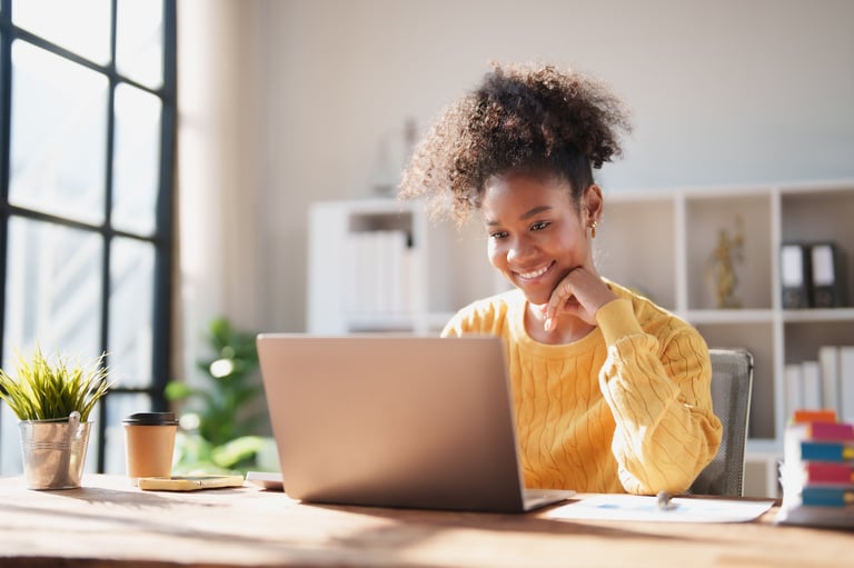 Young African American woman working from a bright home office, smiling at her laptop while studying or managing online business tasks, relaxed and productive in sunlight