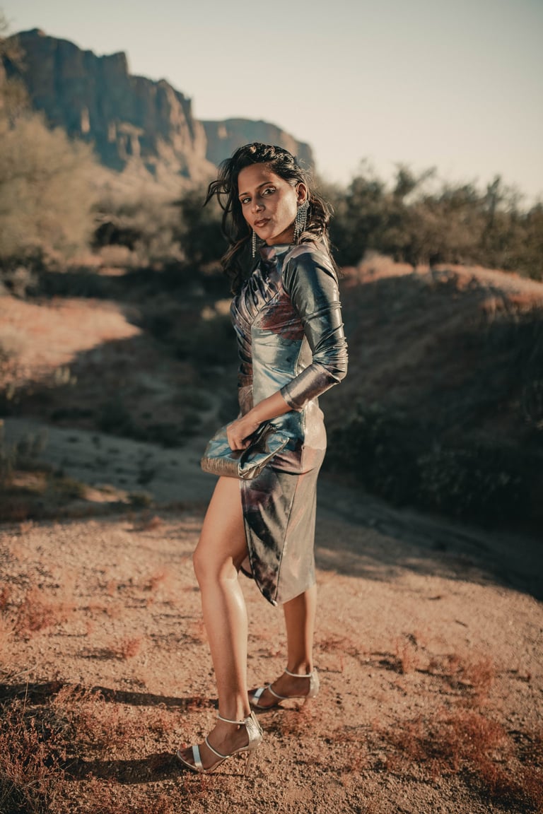 Woman in metallic dress posing on dusty terrain with rocky mountains and desert landscape in background at sunset